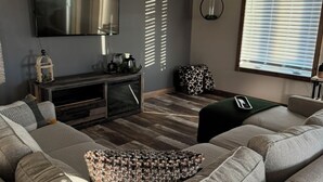 A modern living room with a grey sofa, a flat-screen TV, a ceiling fan, and a window with blinds. - Spacious new home with a country feel. (Kearney)