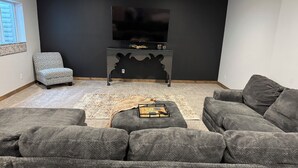 A living room with a large gray sofa, a patterned armchair, a coffee table with remote controls, and a flat-screen TV on a black stand. - Spacious new home with a country feel. (Kearney)