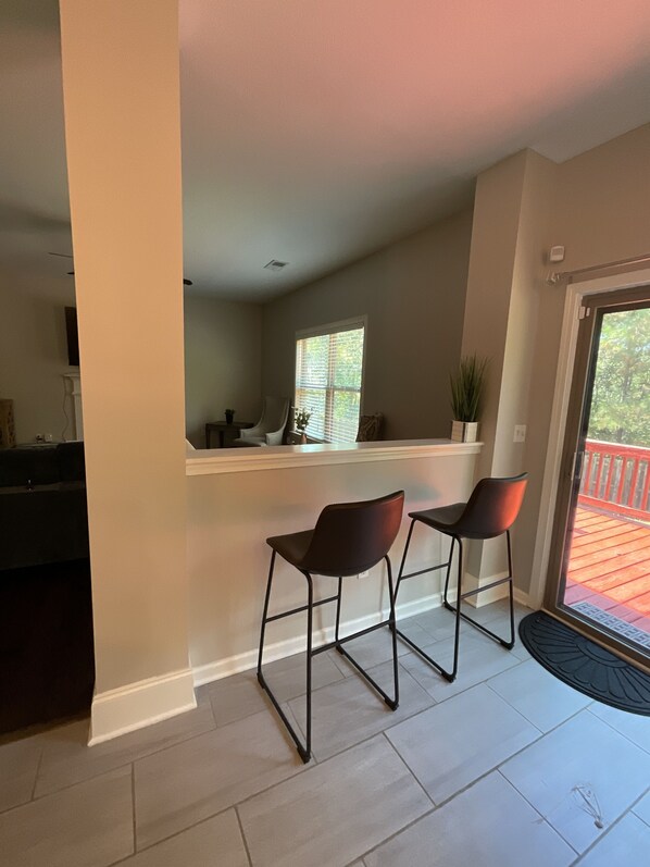 A kitchen with a window, two bar stools, a potted plant, and a sliding glass door leading to an outdoor deck.