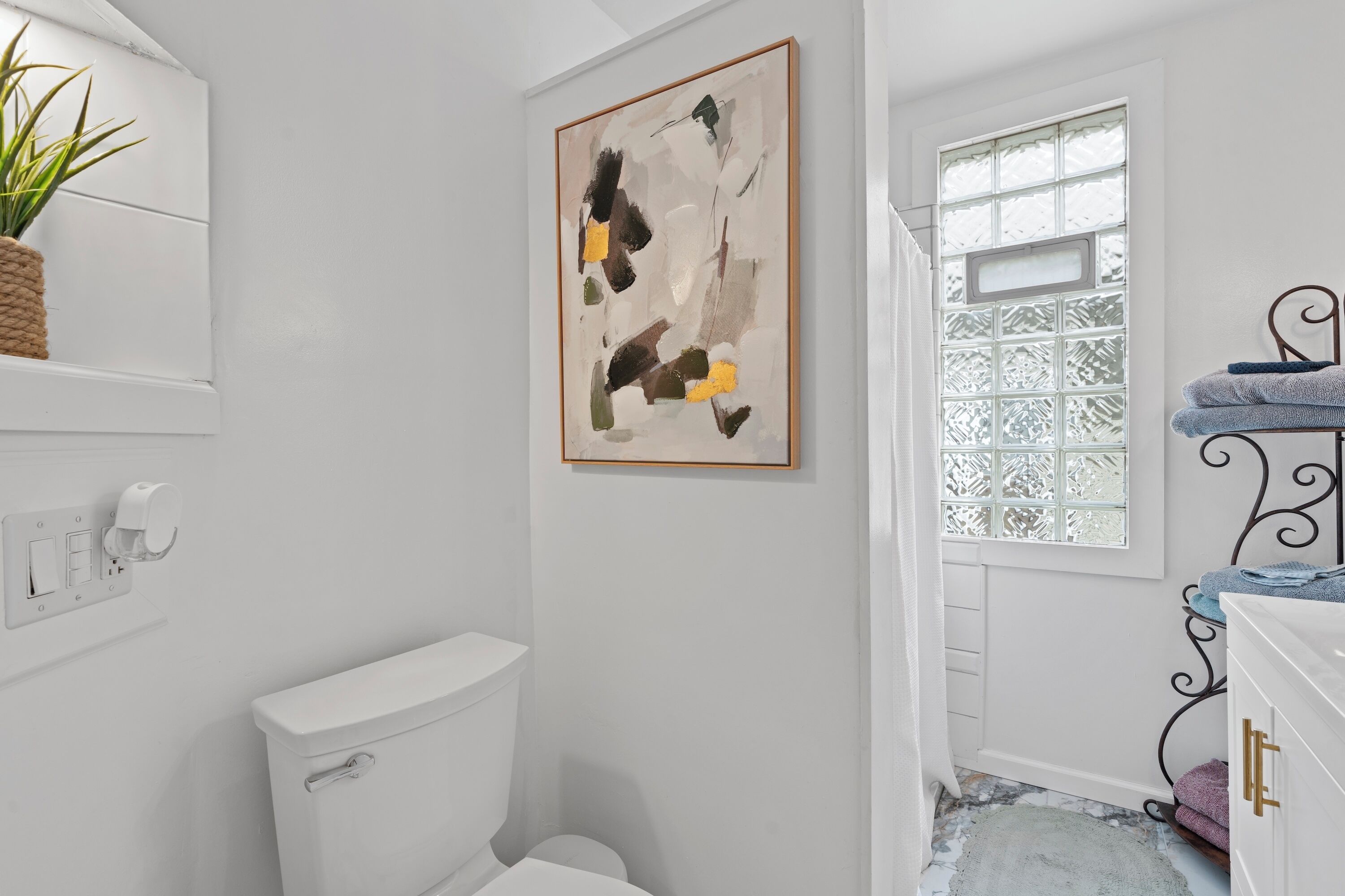 Une salle de bain avec des toilettes blanches, un tableau abstrait encadré, une fenêtre en verre dépoli et un porte-serviettes en métal.