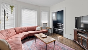 Un salon moderne avec un canapé d’angle rose, une table basse en bois, un tapis à motifs, un fauteuil en cuir noir, un téléviseur posé sur un meuble en bois et une plante suspendue.
