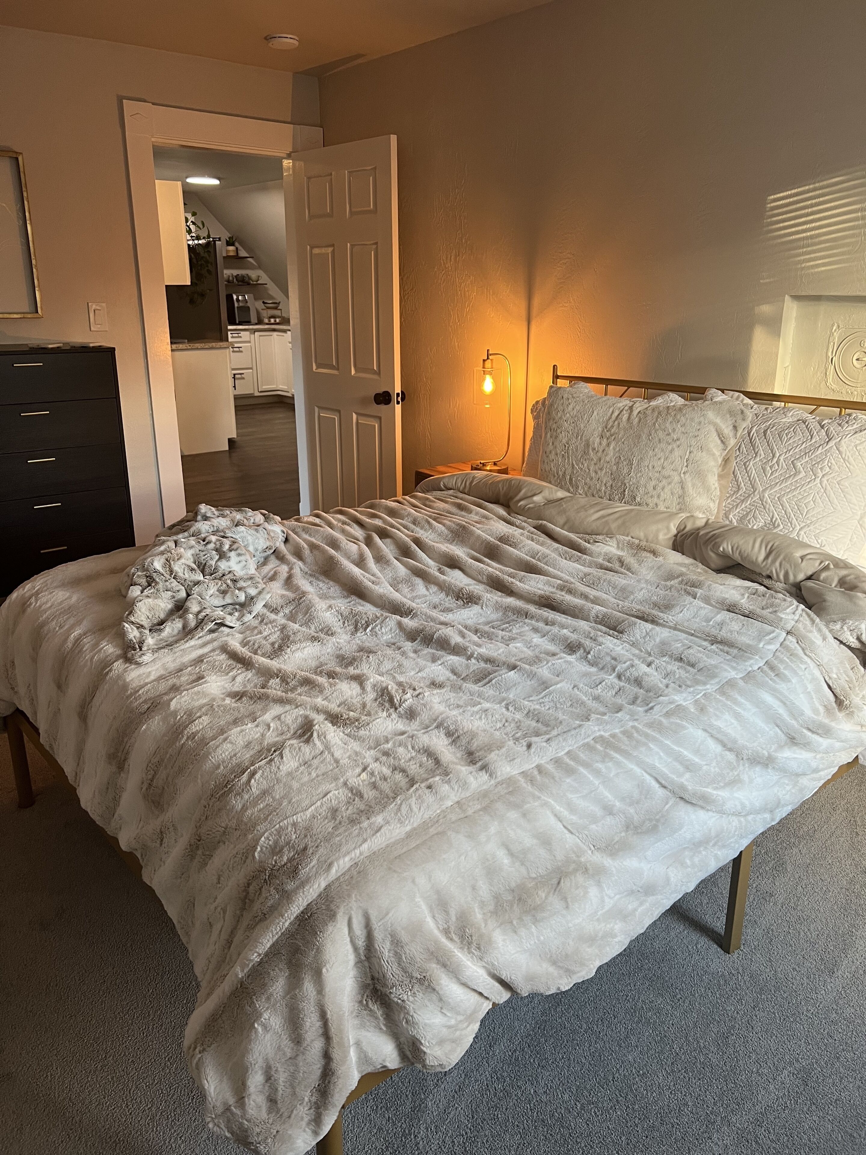 A bedroom with a large bed, a bedside lamp, and a door leading to a kitchen.