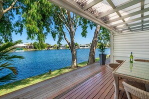 Une terrasse extérieure couverte, dotée d’une table en verre et de chaises en osier, qui offre une vue sur un lac où l’on aperçoit des bateaux et des maisons au loin.