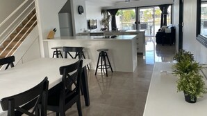 A modern kitchen with a central island, a dining area with chairs, and a staircase leading to another level. - 3 Brm Waterfront Apartment (Noosa)