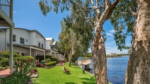A residential area with a house, a boat docked by the water, and a well-maintained lawn. - 3 Brm Waterfront Apartment (Noosa)