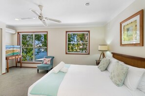 Une chambre d’hôtel avec un lit, deux fenêtres, un bureau avec un tableau, une chaise et un ventilateur de plafond.