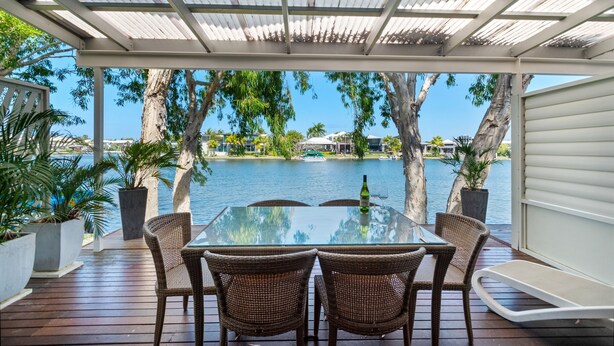 A covered outdoor dining area with a glass table and wicker chairs, overlooking a body of water and trees. - 3 Brm Waterfront Apartment (Noosa)