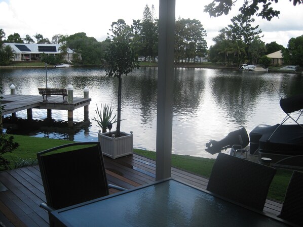 Une terrasse avec une table et des chaises donnant sur un lac paisible, entourée de verdure et avec quelques bâtiments visibles au loin.