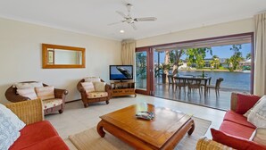 A living room with a television, wicker furniture, a red sofa, and a view of a body of water through large glass doors. - 3 Brm Waterfront Apartment (Noosa)