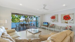 Un salon spacieux doté d’une grande fenêtre donnant sur une piscine, d’un ventilateur de plafond et d’un téléviseur à écran plat posé sur une table en bois.