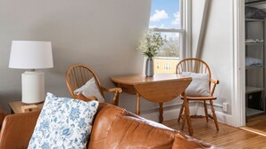 Una habitación acogedora con una mesa de madera, dos sillas de madera, una lámpara blanca, un sofá con un cojín estampado y una ventana con vistas a edificios.