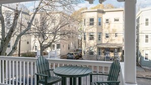 Un porche cubierto con una mesa redonda y dos sillas, con vista a una calle con coches aparcados y un edificio de varios pisos.