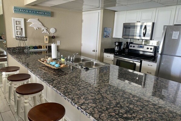 A kitchen with granite countertops, white cabinets, a stainless steel refrigerator, and a microwave. There are bar stools along the counter.