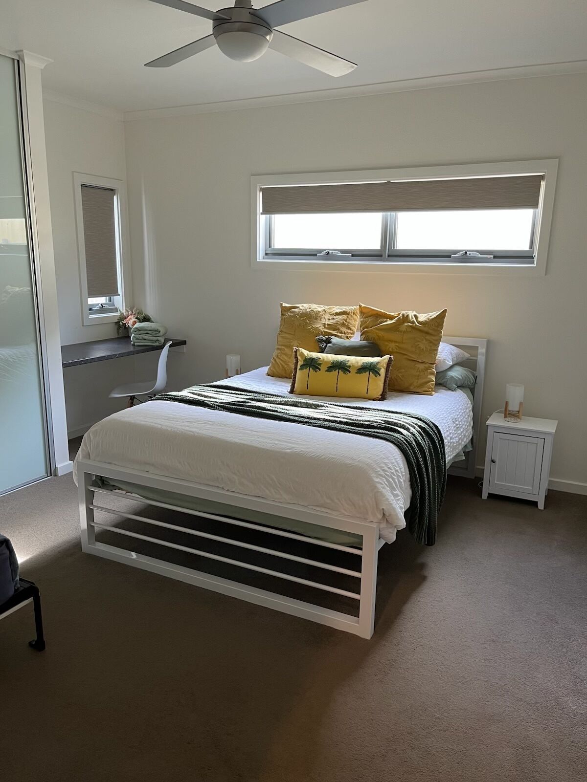 A bedroom with a bed, a ceiling fan, a window with blinds, a nightstand, and a mirror.