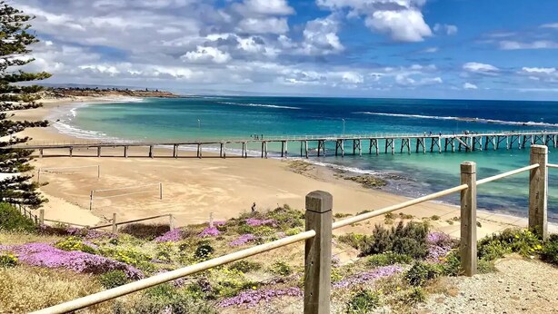 Une plage avec une jetée en bois qui s’avance dans l’océan, entourée d’un ciel dégagé et de quelques nuages épars.
