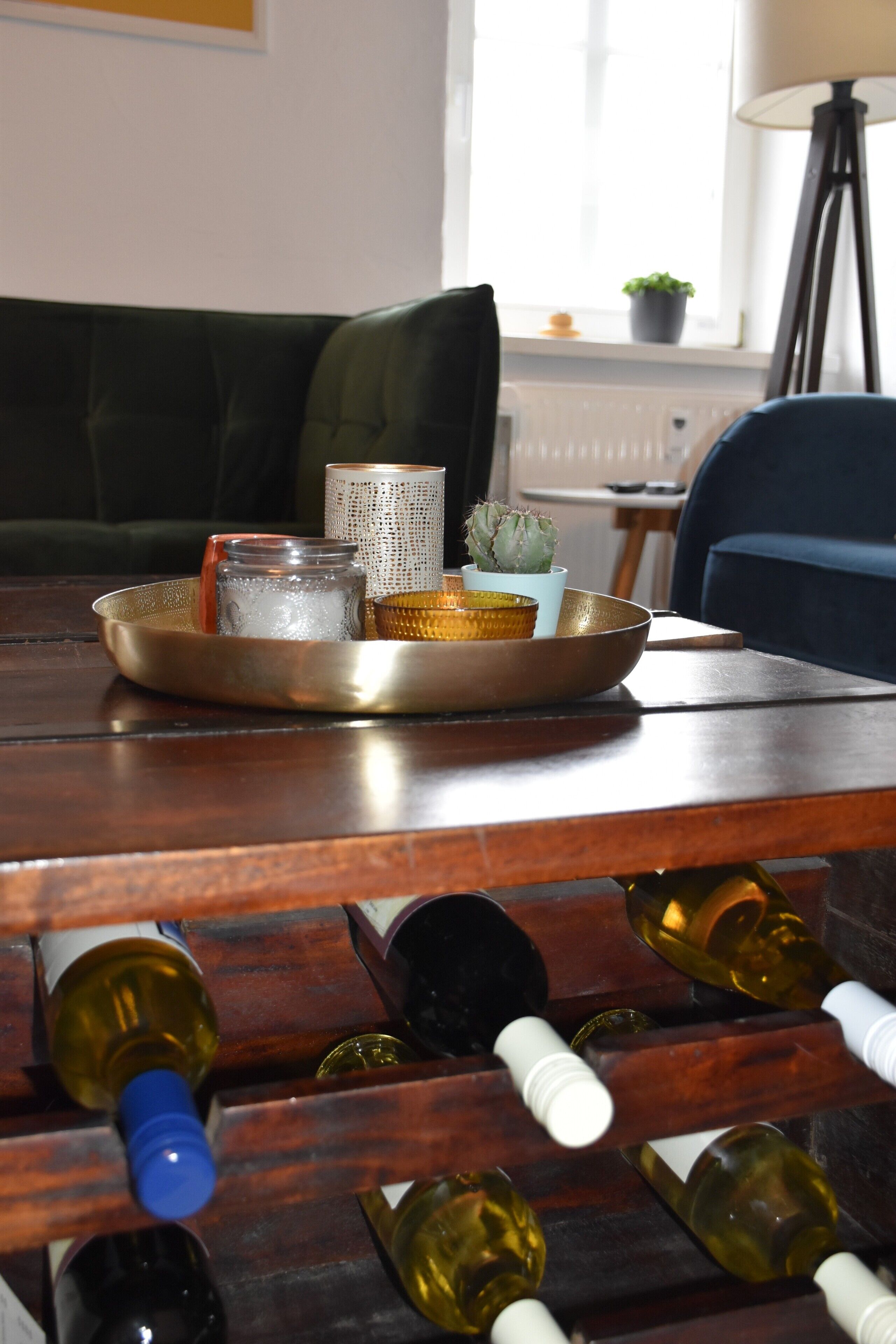 A wooden table with a tray holding a glass jar, a small potted plant, and a woven container. Below the table, there are wine bottles arranged in a rack.
