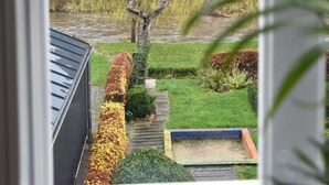 Vista desde una ventana que muestra un jardín con un banco de madera, un estanque pequeño y una colina con árboles al fondo.