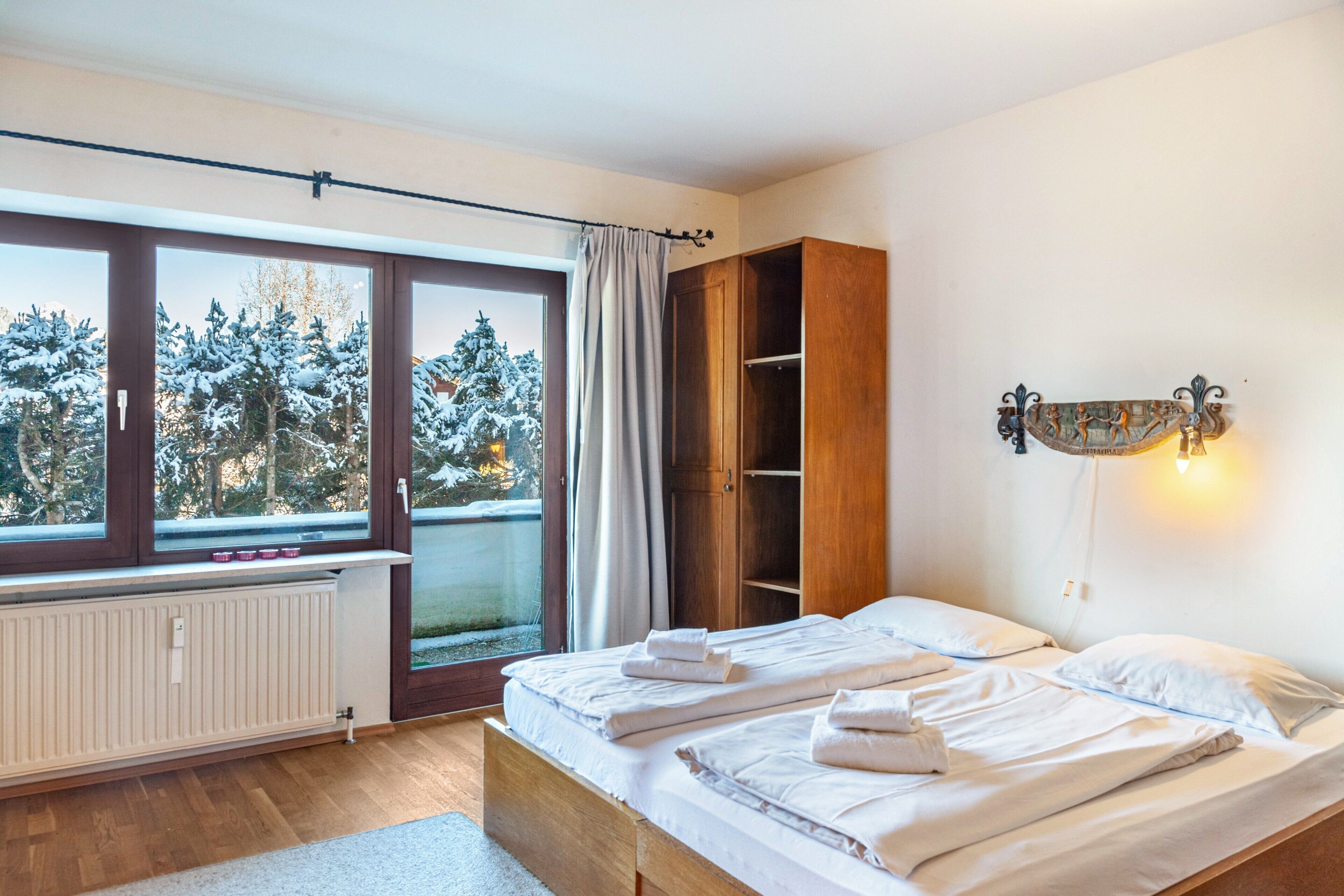 A bedroom with a large window, a radiator, a wooden wardrobe, and a wall-mounted light fixture.