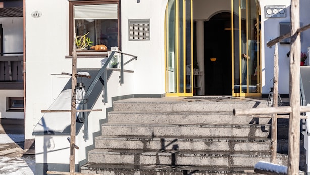 L’entrée d’un hôtel, avec une enseigne portant l’inscription « EXCELSIOR », un escalier menant à l’entrée et une fenêtre avec des stores.