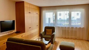 Un salon comprenant un téléviseur, des meubles en bois, un canapé, un pouf et un fauteuil.