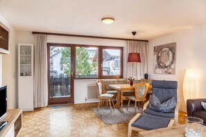 Une pièce avec un plancher en bois, une table à manger avec des chaises, un canapé, un téléviseur, une fenêtre avec des rideaux et un radiateur.