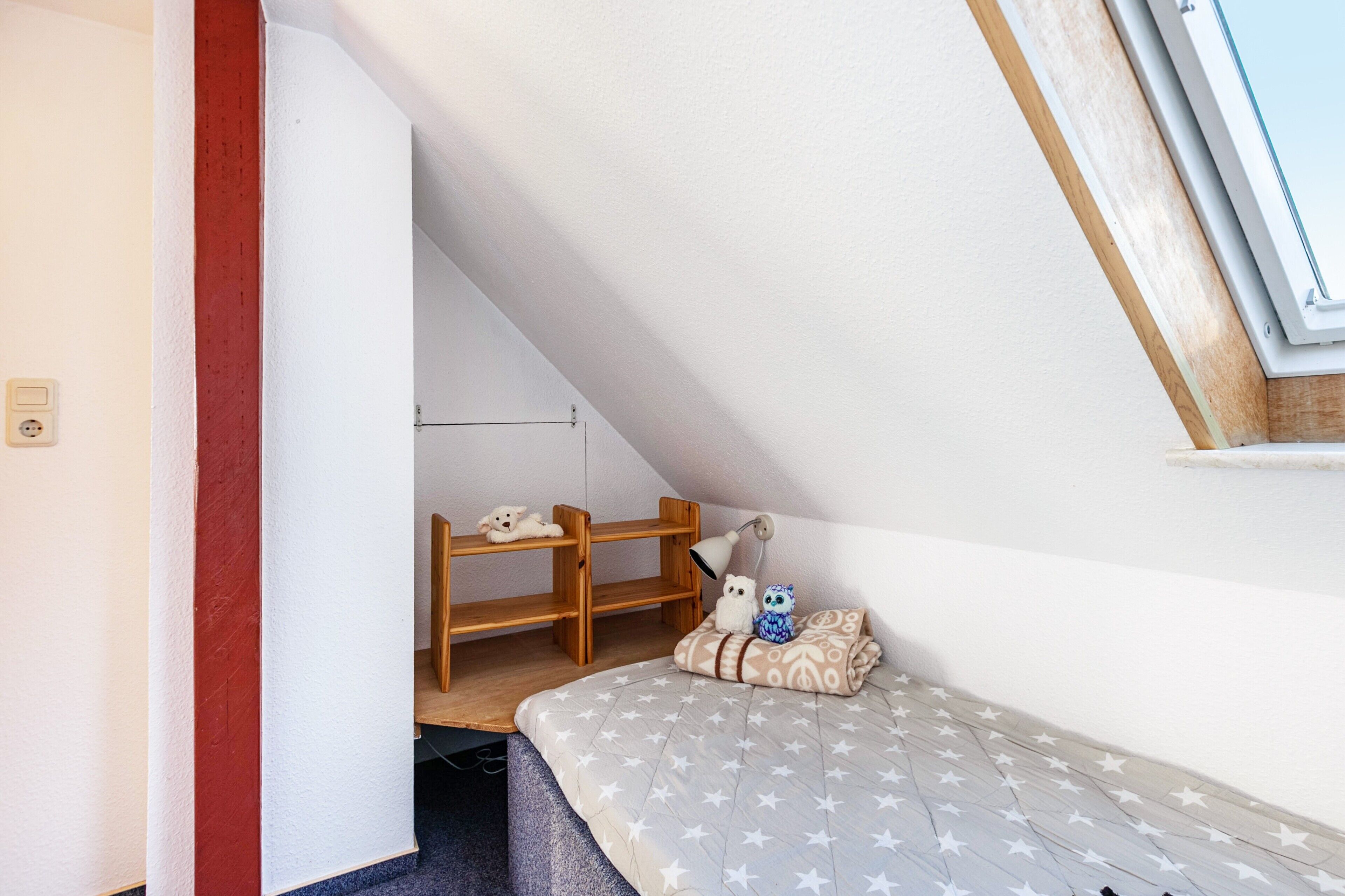 Une petite pièce chaleureuse avec un plafond en pente, un lit recouvert d’une couverture à motifs d’étoiles, une étagère en bois remplie de jouets et une fenêtre avec un cadre en bois.