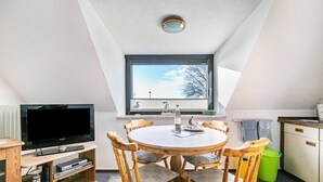 A room with a round wooden table, six chairs, a television on a wooden stand, a window with blinds, and a green trash bin. - Apartment "Whg 2 - Vacation on a Country Estate" with Sea View, Shared Garden, and Wi-Fi (Schönhagen)