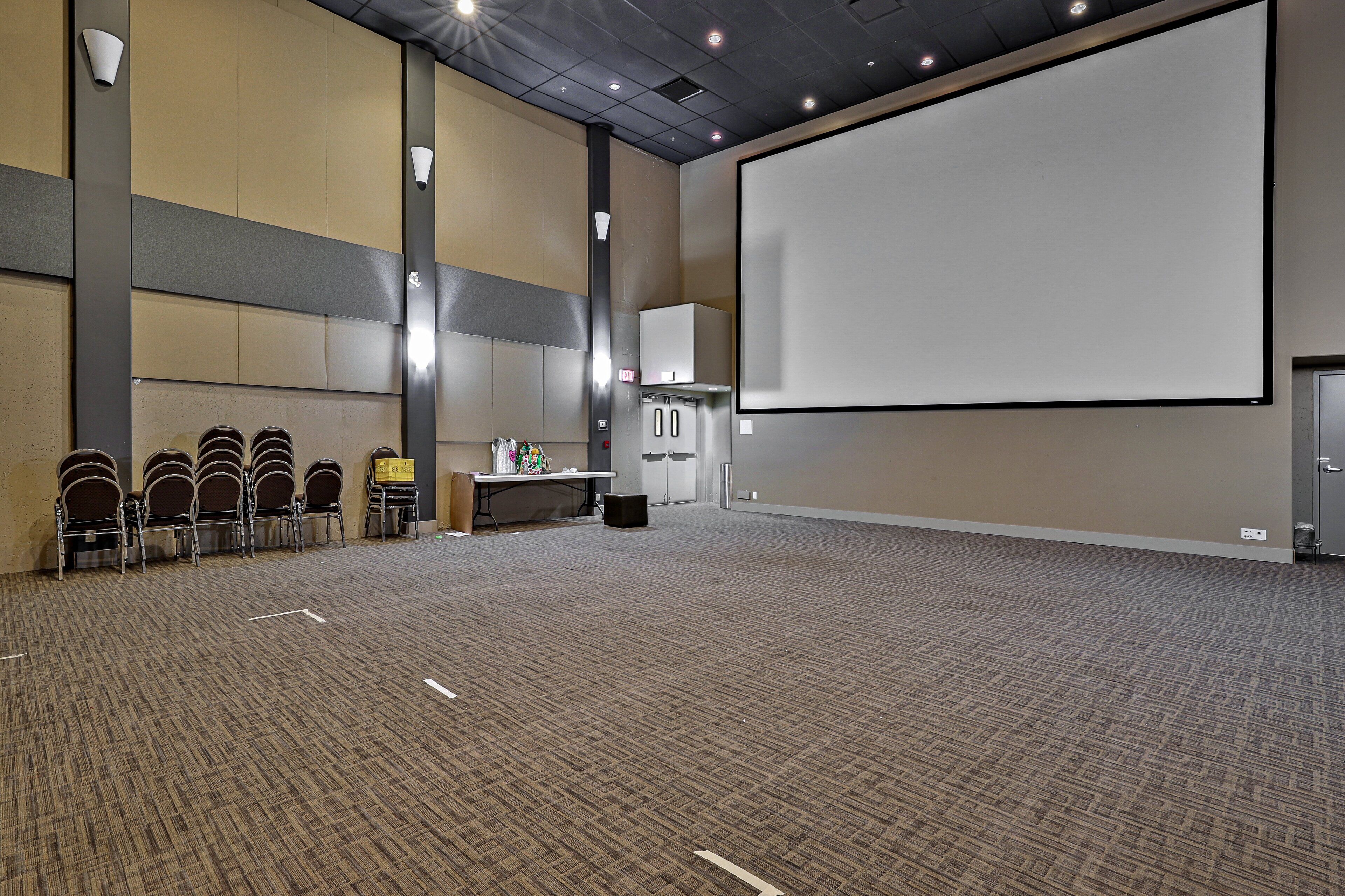 Une grande salle de conférence dotée d’un sol recouvert de moquette, d’un écran de projection et de plusieurs rangées de chaises.