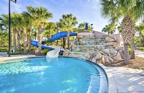 A pool area with a water slide, palm trees, and a rocky waterfall backdrop. - Davenport/Orlando Vacation House, Game Room, Water Park Access, Heated Pool. (Davenport)
