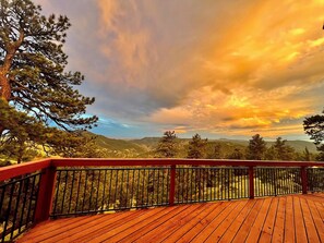 Una terraza de madera con barandilla de metal, con vistas a un paisaje montañoso con árboles y un cielo al atardecer.