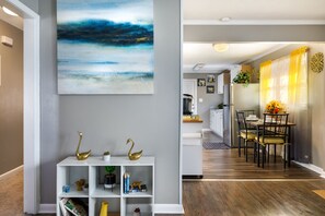 A modern living room with a white console table, a large abstract painting, and a dining area with a wooden table and chairs. - This is a cozy vintage ranch that sits on the edge of HWY 95 boarders.  (Fayetteville)