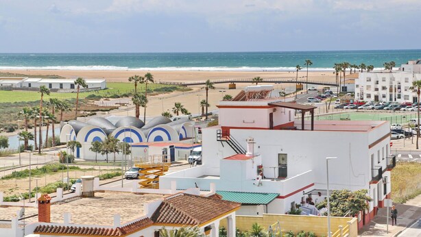 A coastal area with a beach, palm trees, and buildings including a white structure with a red roof and a blue and white building with circular designs. - Ceuta apartment near the beach (Conil de la Frontera)