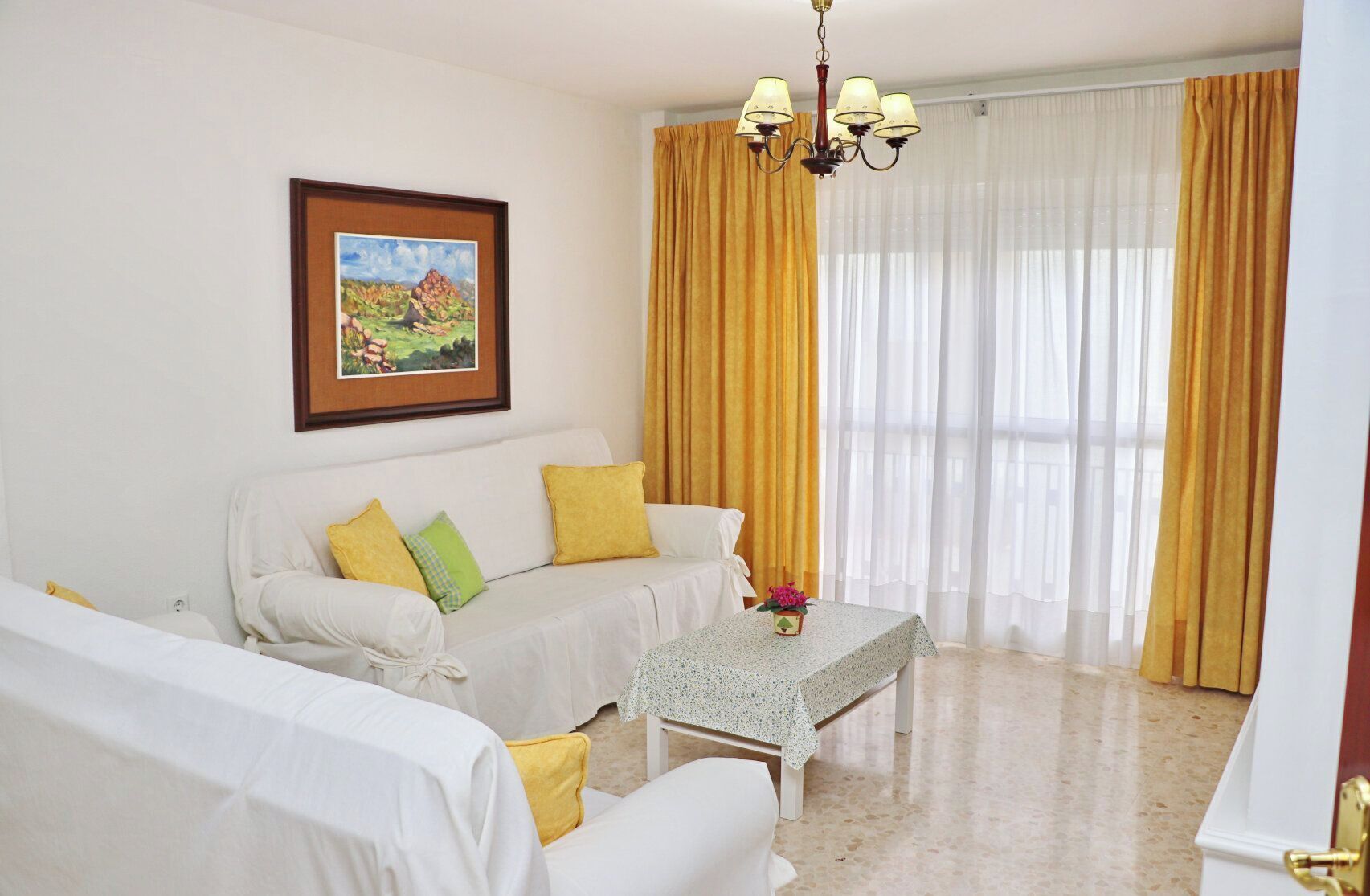 A room with a white sofa set, a small table with a vase, a chandelier, and a painting on the wall.