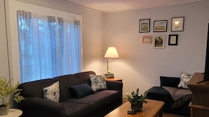 Uma sala de estar com um sofá escuro, uma mesa de centro de madeira e um abajur. Há quadros nas paredes e uma janela com cortinas.