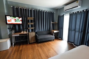 A modern hotel room with a wooden floor, a grey sofa, a desk with a computer, a TV, and an air conditioner. - A Hotel Cozy (Chiang Saen)