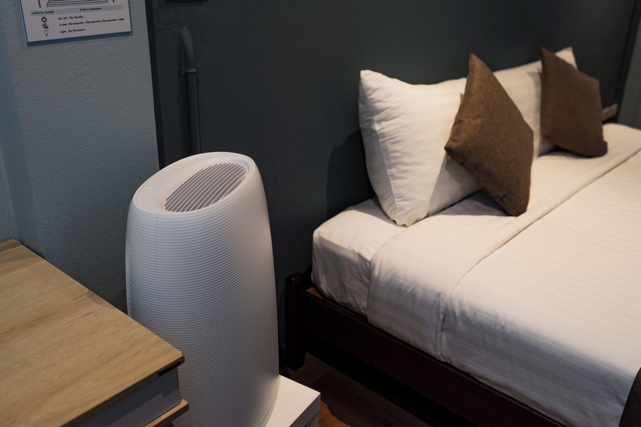 A hotel room with a bed, pillows, a white air purifier, and a wooden nightstand.