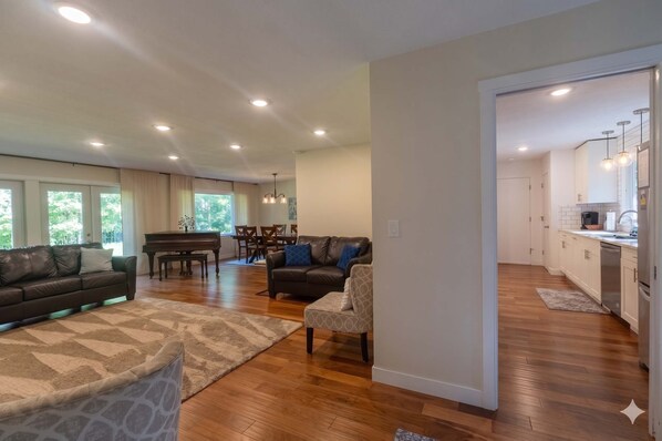 A spacious living area with a sofa, armchair, and piano, leading to a dining area with a table and chairs.