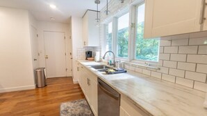 A kitchen with white cabinets, a stainless steel sink, and a window with a view of greenery. - Stunning Ranch in the woods! (Berrien Springs)