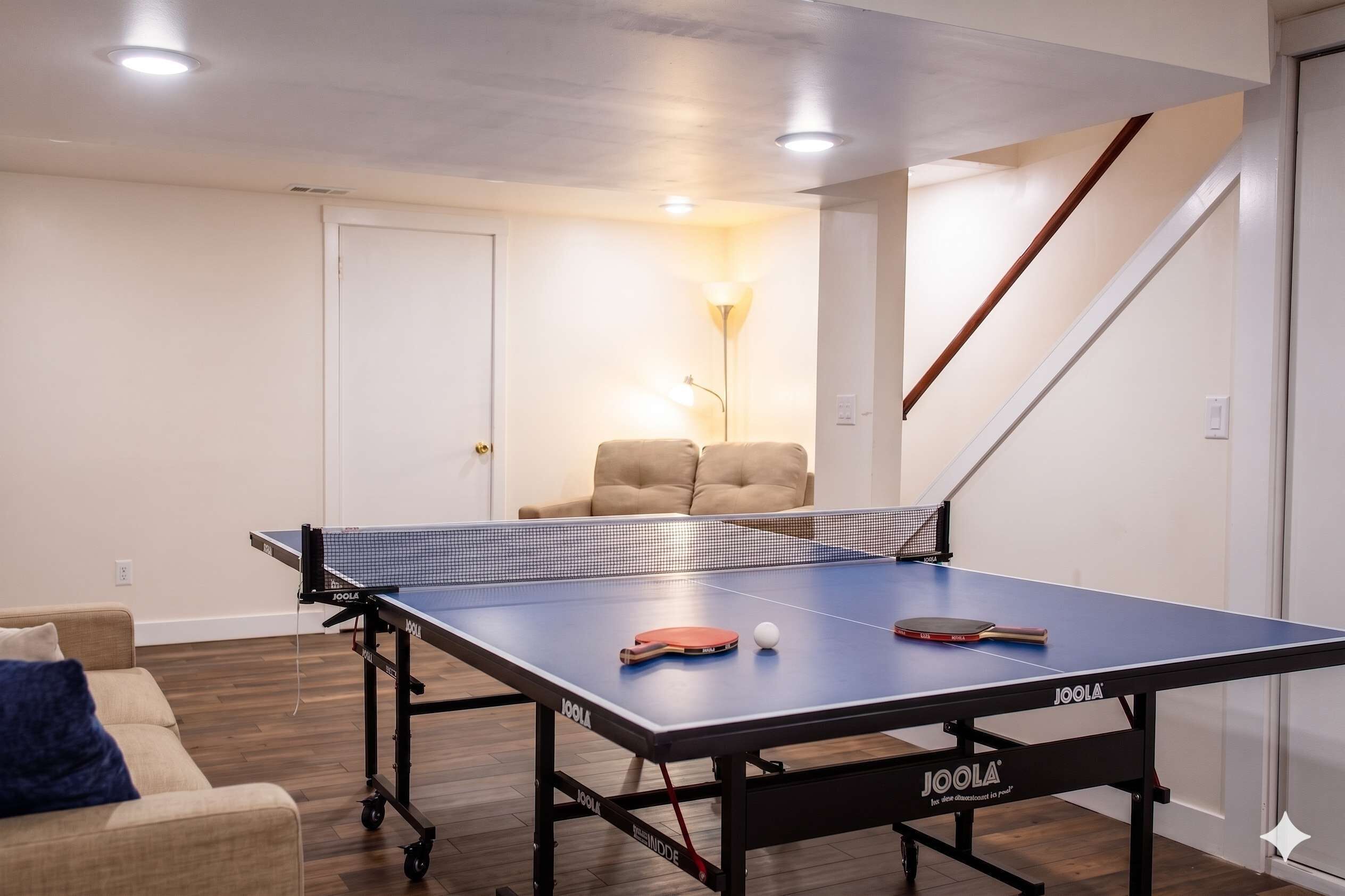 Una mesa de ping pong con red, una paleta de tenis de mesa y una pelota en una habitación que tiene un sofá, una lámpara y una escalera.