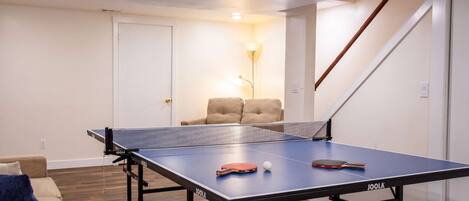 Una mesa de ping pong con red, una paleta de tenis de mesa y una pelota en una habitación que tiene un sofá, una lámpara y una escalera.
