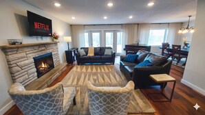 A living room with a stone fireplace, a black leather sofa, a patterned armchair, a dining area with a piano, and a TV displaying 'NETFLIX'. - Stunning Ranch in the woods! (Berrien Springs)