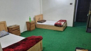A room with two wooden beds, green carpet, a small wooden nightstand with a bottle, and a door in the background. - Sada Bahar Inn Hotel (Gilgit)