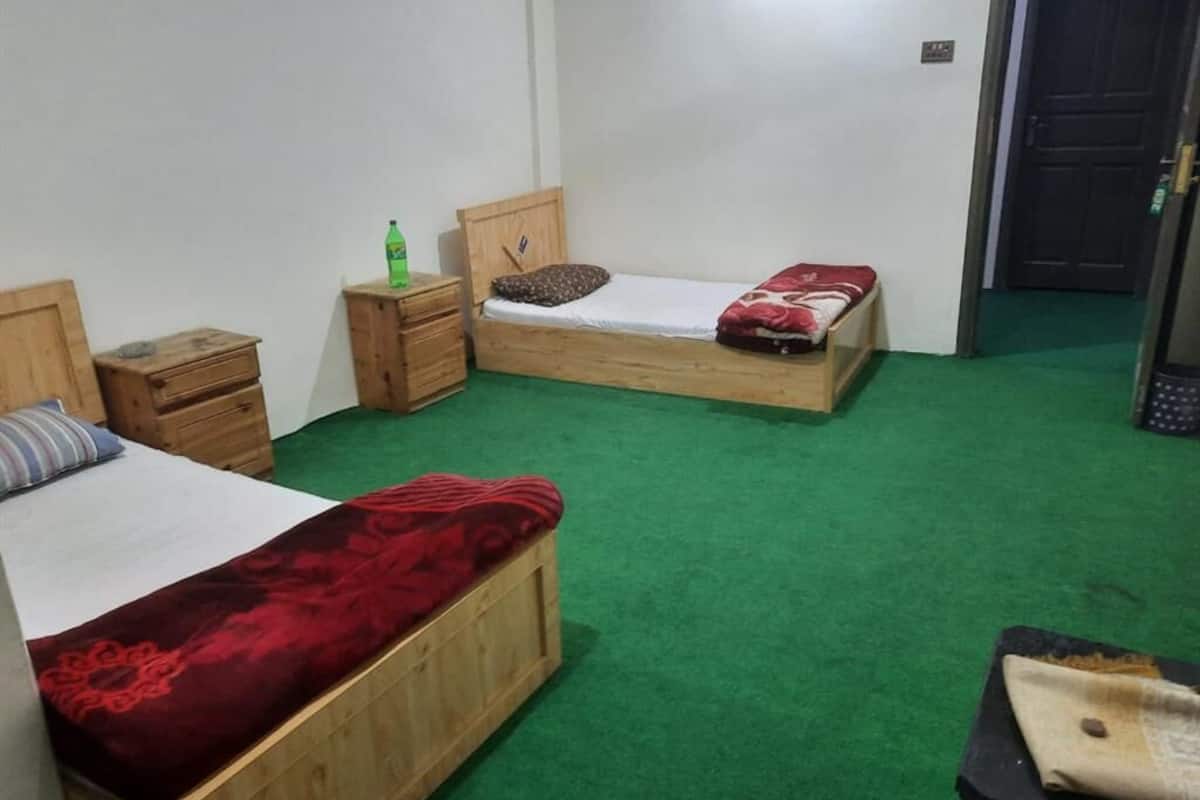A room with two wooden beds, green carpet, a small wooden nightstand with a bottle, and a door in the background.