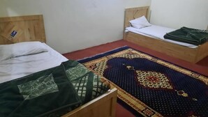 Two wooden beds with white linens and a patterned rug in a simple room with plain walls. - Sada Bahar Inn Hotel (Gilgit)
