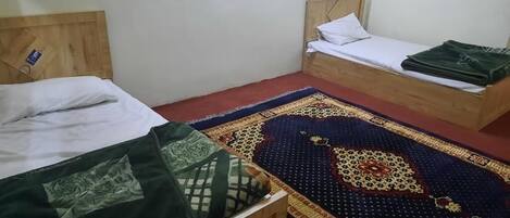 Two wooden beds with white linens and a patterned rug in a simple room with plain walls.
