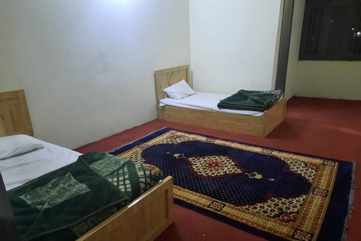 A room with two wooden beds, one with a green blanket and the other with a white pillow. A colorful rug is on the floor.