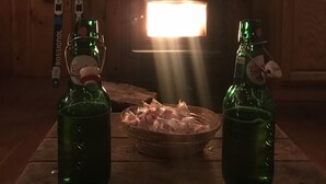 Een houten interieur met een tafel waarop twee groene flessen, een mand met eten en een open haard staan die licht geeft.