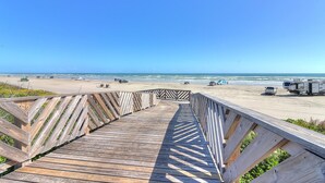Un paseo marítimo de madera que conduce a una playa, con vehículos y un cielo azul despejado.