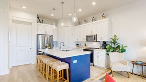 Una cocina moderna con armarios blancos, una isla central con taburetes, electrodomésticos de acero inoxidable, lámparas colgantes y una mesita auxiliar con una planta.