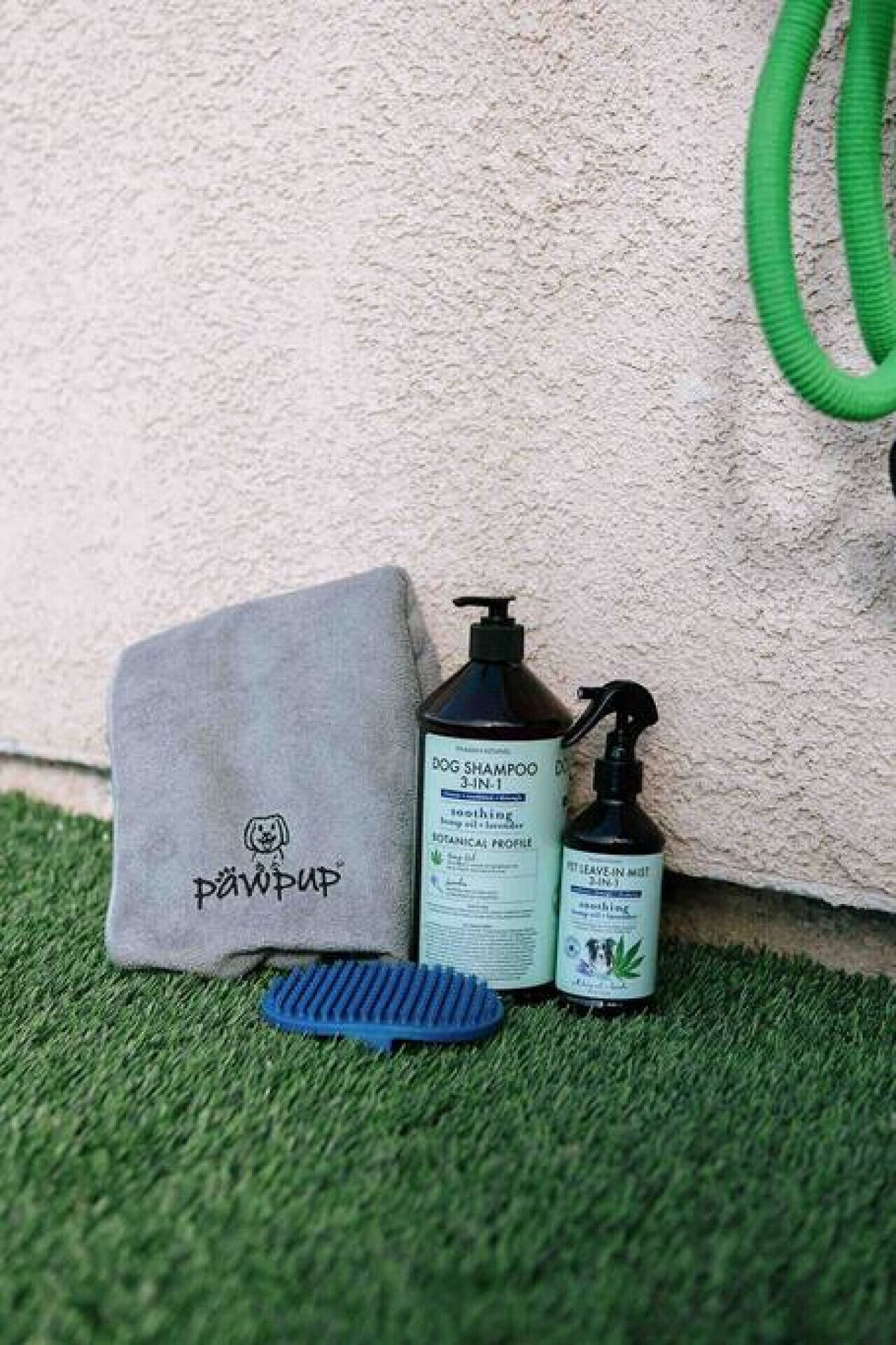 Un sac gris orné d’un logo, deux bouteilles de shampoing pour chiens et une brosse bleue, le tout posé sur une surface verte.